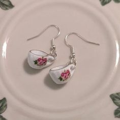 two tea cups with pink flowers on them are sitting on a white plate, next to silver earwires