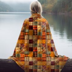 a woman is sitting on a log looking out at the water while wearing a plaid blanket