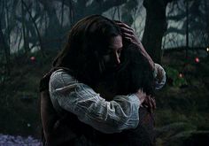 a man and woman hug in the woods at night, with lights on trees behind them