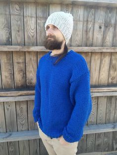 a man with a beard wearing a blue sweater and tan pants standing in front of a wooden fence