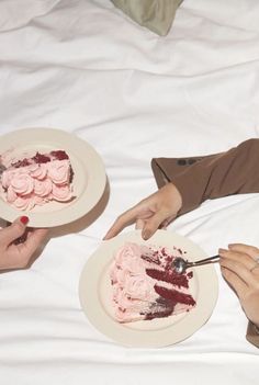 two people sitting on a bed holding plates with pink frosting and one person cutting the cake