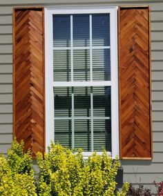 a window with wooden shutters on the side of a gray house next to shrubbery