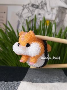 an orange and white beaded hamster sitting on top of a wooden stick next to some green grass