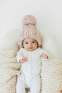 a baby wearing a knitted hat laying on top of a white blanket and pillows