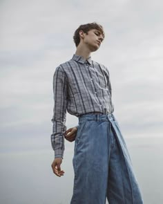 a man standing on top of a beach next to the ocean wearing blue jeans and a plaid shirt