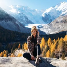 a woman sitting on top of a rock with mountains in the background and trees around her