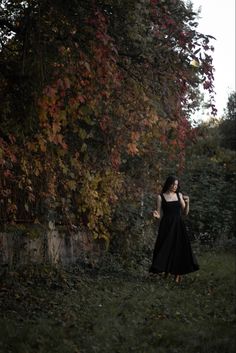 a woman in a long black dress is standing near a tree with red and yellow leaves