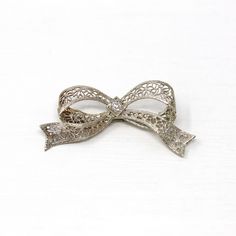 Gorgeous vintage circa 1930s era 14k white gold diamond bow brooch! This beautiful piece showcases a sparkly old European cut diamond center weighing an estimated .07 carats. The setting features ornate flower filigree with milgrain details. The brooch secures with a rotating c-clasp. An outstanding Art Deco Era piece of fine jewelry! *Sale - price reduced from $340 USD to $330 USD.  ERA - Circa 1930s - Art Deco  METAL / MATERIAL - 14k white gold, 1 old European cut diamond (estimated .07 cts, SI2, near colorless)  MARKINGS / HISTORY - Brooch clasp marked "14k"  CONDITION - Good antique condition. White gold metal has been professionally polished & cleaned. Very light age appropriate patina & wear remains. Amazing vintage bow brooch!  SIZE / MEASUREMENTS - Brooch: 1 1/2 x 3/4 inches, Weigh Elegant Filigree Brooches For Evening, Elegant Brooches With Intricate Design For Gift, Elegant Brooches With Intricate Design For Anniversary, Elegant Platinum Brooches For Gift, Elegant Anniversary Brooch With Bow, Silver Brooch With Decorative Bow, Silver Brooches With Decorative Bow, Vintage Platinum Brooches For Anniversary, Vintage Platinum Brooches For Wedding