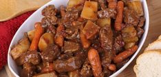 a white bowl filled with meat and carrots on top of a wooden table next to bread