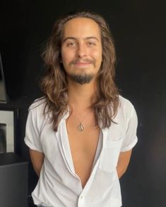 a man with long hair and no shirt standing in front of a black wall smiling at the camera