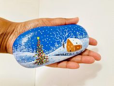 a hand holding a painted rock with a house on it and a christmas tree in the snow