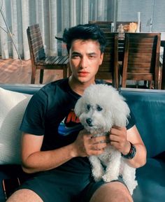 a man sitting on a couch holding a small white dog in his lap and looking at the camera