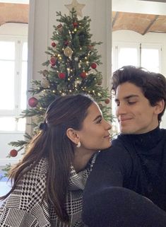 a young man and woman sitting next to each other in front of a christmas tree