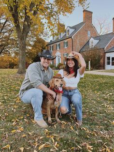 a man and woman pose with their dog in front of a large brick house on a fall day