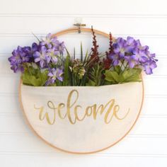 a welcome sign hanging on the wall with purple flowers in it and greenery inside