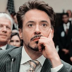 the man is wearing a suit and tie while sitting in front of an audience with his hand on his chin