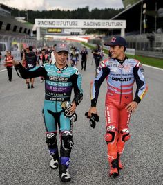 two men walking down a race track with helmets on and one holding his hand in the other's pocket