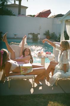 three women sitting on lounge chairs in front of a swimming pool, one with her arms up