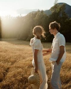 two people standing in the middle of a field with grass and trees behind them, looking at each other