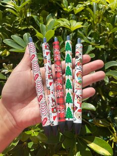 a hand holding four pens with christmas designs on them and trees in the back ground