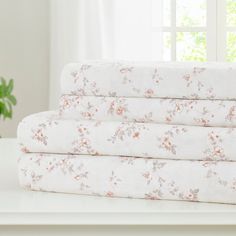 a stack of white sheets with pink flowers on them sitting on a table next to a potted plant