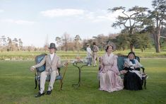 three people sitting on chairs in the grass with one woman holding a cane and another man wearing a top hat