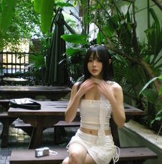 a woman sitting on top of a wooden bench in front of green plants and trees