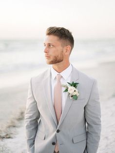 a man in a gray suit and pink tie standing on the beach with his hands in his pockets