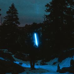 a man standing in the snow at night with a light on his back and trees behind him