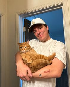 a man holding a cat in his arms and smiling at the camera while wearing a baseball cap