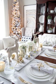 a dining room table set for christmas with white and gold decorations, silverware and deer figurines