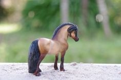 a small toy horse standing on top of a sandy ground next to grass and trees