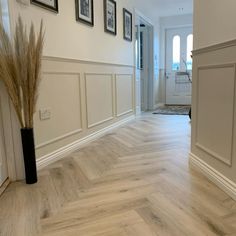 an empty hallway with white walls and wood flooring on either side is a black vase with dry grass in it