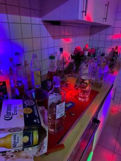 a kitchen counter covered in liquor bottles and empty glasses on it's side, next to a neon light