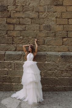 a woman in a white dress leaning against a brick wall with her arms up and hands behind her head