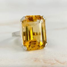 Vibrant emerald cut citrine in a solid and heavy basket setting. The citrine has a small black inclusion off to the side and I like to think of it as a birthmark. In very good condition with some light wear to the citrine and setting.  11.5 carat citrine that measures 16.9mm x 11.9mm. Ring sits 9.6mm high off the finger.  14k white gold, size 4 and sizable. Weighs 7.55 grams.  Free shipping on all domestic orders via USPS First Class insured with signature confirmation. Shipping upgrades available at checkout.  We offer international shipping. Please contact us for details. Most of our pieces are vintage or antique and therefore are not in perfect condition. Please keep this in mind when purchasing. It should be expected that these pieces will have normal wear and tear according to their a Yellow Emerald-cut Topaz Ring, Yellow Citrine Topaz Ring Rectangular Shape, Classic Emerald Cut Yellow Topaz Ring, Classic Yellow Emerald Cut Topaz Ring, Classic Yellow Topaz Ring With Emerald Cut, Yellow Topaz Ring With Emerald Cut And Prong Setting, Yellow Topaz Ring With Prong Setting, Emerald Cut, Yellow Emerald Cut Topaz Ring With Prong Setting, Classic Yellow Rectangular Rings