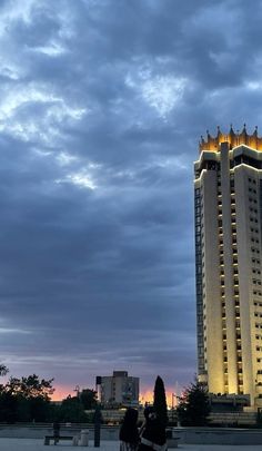 a woman sitting on a bench in front of a tall building with lights at the top