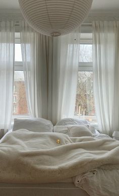 an unmade bed in front of two windows with drapes hanging from the ceiling