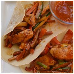 two tacos with chicken, peppers and onions next to a small bowl of sauce