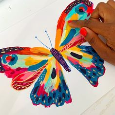 a hand holding a colorful butterfly on top of a white sheet with watercolors