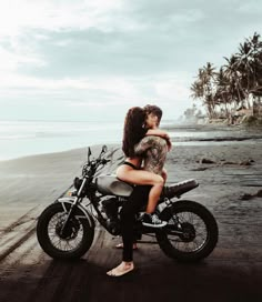 two people sitting on a motorcycle by the beach with palm trees and water in the background