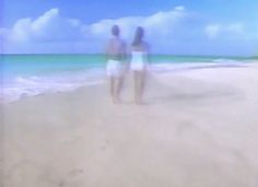 two people are walking on the beach in front of the blue sky and ocean water