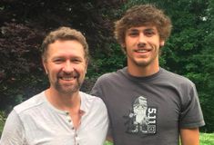 two men standing next to each other in front of trees and bushes, one smiling at the camera