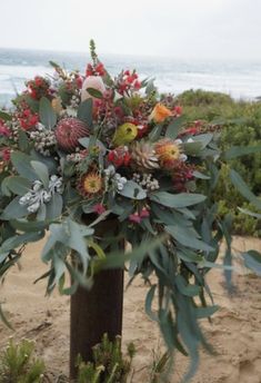 an arrangement of flowers and greenery in a vase on the sand near the ocean