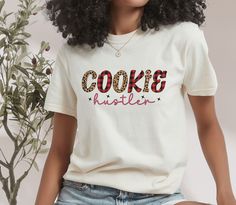 "Indulge in the sweet side of life with our \"Leopard & Buffalo Plaid 'Cookie Hustler' T-Shirt,\" a playful and stylish choice for the baker who knows their way around a cookie sheet. With its unique blend of patterns and sassy script, this tee is perfect for those who hustle as hard in the kitchen as they do in life. The unisex soft-style t-shirt puts a new spin on casual comfort. Made from very soft materials, this tee is 100% cotton for solid colors. Heather colors and sports grey include pol Cookie Brand Shirt, Cookie Shirts Ideas, Funny Baking Shirts, Funny Baking, Cookie Shirt, Girl Scout Cookies, Girl Scout, Cookie Sheet, Soft Style