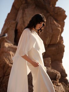 a woman standing in front of some rocks wearing a white cape and wide leg pants