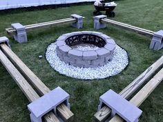 an outdoor fire pit surrounded by benches in the middle of some grass with a man sitting on a lawn mower behind it