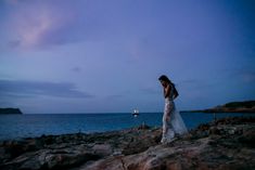 a woman standing on top of a rocky beach next to the ocean at night with her dress blowing in the wind