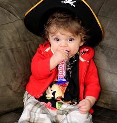 a small child sitting on top of a couch wearing a pirate hat and eating a candy bar