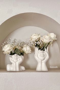 two white vases with flowers in them on a shelf